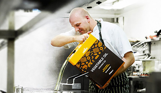 A chef pours liquid from an orange and black box into a vat in a kitchen