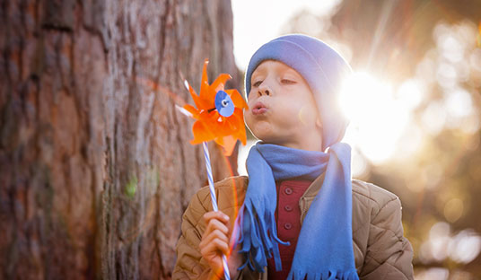 A young boy wearing a blue hat and scarf blows on a small orange toy windmill in a forest