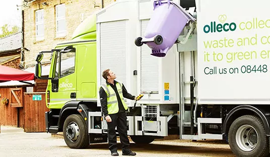 A garbage truck lifts a purple bin as a bin man looks on